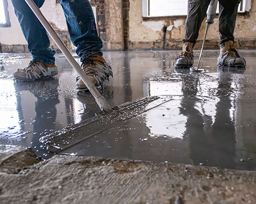 Contractors smoothing a freshly poured self-leveling gypsum underlayment on a subfloor using a gauge rake.